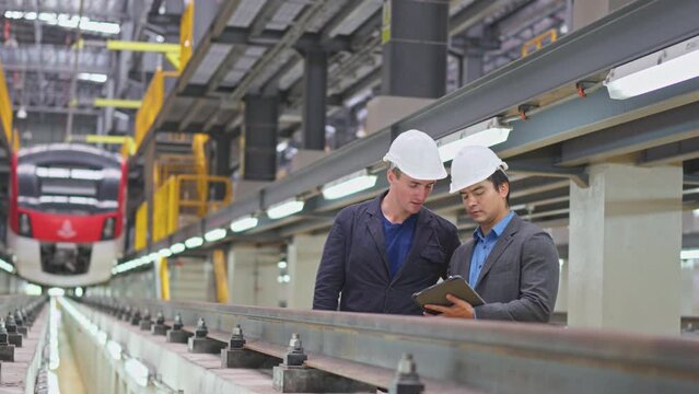 Front view of professional manager or engineer workers discuss together with tablet and stand near the railroad tracks of electrical or metro train in the factory.