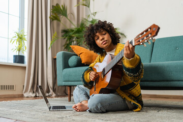 African American woman learning guitar watching tutorial on laptop at home. Black girl playing guitar singing song learning online music lessons. Artistic woman playing acoustic guitar learning chords