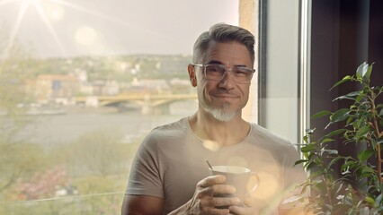 Casual older man standing by window at at home drinking morning coffee. Portrait of happy mid adult, middle aged man in 50s, smiling. Cityscape with river and bridge in background.