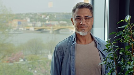 Casual older man standing by window at at home. Portrait of happy mid adult, middle aged male in 50s, smiling. Cityscape with river and bridge in background.