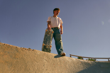 Low angle of happy male skateboarder holding skateboard standing at skate park in sunny day - obrazy, fototapety, plakaty