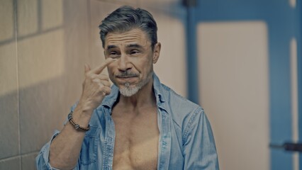 Mature adult man in open shirt applying cosmetics in bathroom mirror. Using anti-aging eye cream...