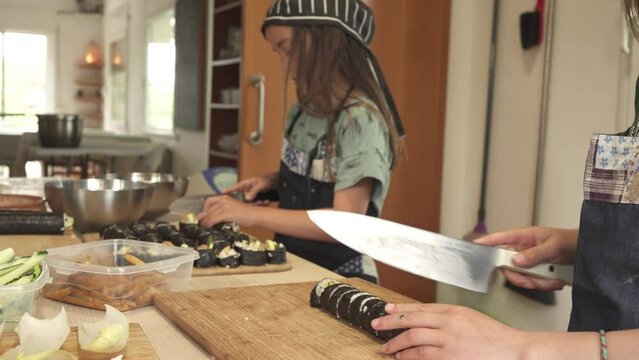 nine-year-old twins cutting sushi rolls with knives, wearing aprons and chef's hats, concentrating on rolling nori seaweed with rice, tofu and avocado.