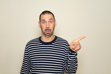 Hispanic man with beard in his 40s wearing a striped sweater pointing to the side with finger with a surprised face, isolated on beige background.