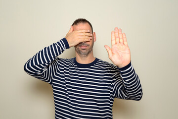 Hispanic man with beard in his 40s wearing a striped sweater covering his eyes with his hands and making stop gesture with a sad and afraid expression. Embarrassed and negative concept