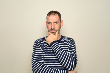 Hispanic man with beard about 40 years old wearing striped sweater thinking concentrated in doubt with finger on chin and looking at the camera wondering, isolated over beige background.