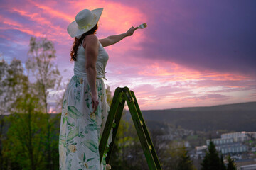 a woman in a dress stands on a wooden ladder and paints the sunset