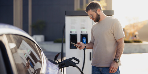 A man plugs his electric vehicle into a charging station while looking at his smartphone on a sunny...