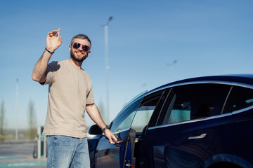 Smiling bearded man with sunglasses charges his modern electric vehicle at a charging station under a clear blue sky, showcasing environmental consciousness and sustainable living.