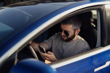 Smiling, bearded man wearing sunglasses interacts with his smartphone while sitting in the driver's seat of a modern electric car, showcasing a blend of technology and eco-friendly transportation.
