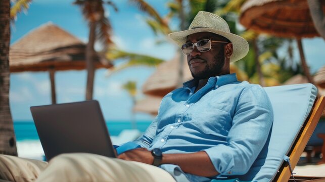 A focused man in a blue shirt working on his laptop while relaxing on a beach chair under a sunny sky