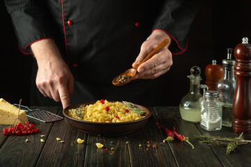 The chef adds aromatic spices or dry pepper with a spoon to a bowl of pasta on the kitchen table. Low key concept of preparing a delicious breakfast in the cook room