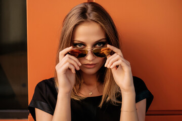 girl standing by an orange wall and adjusting her sunglasses
