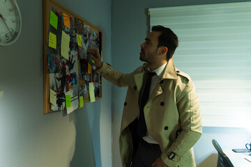 Male detective in trench coat reviews evidence on a corkboard in a shadowy workspace