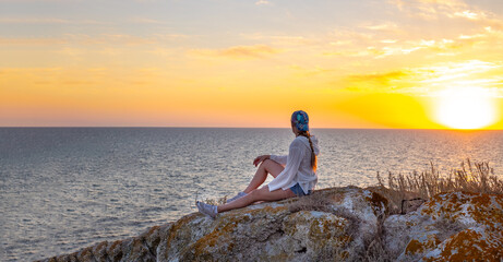 A traveler sits on a mountain top on the seashore and looks at the sunset sky.