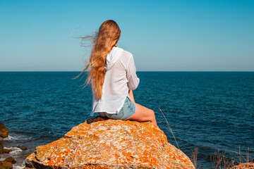 A woman with long blond hair sits on a rock on top of a cliff and looks out at the dark blue sea. Travel and tourism in summer.