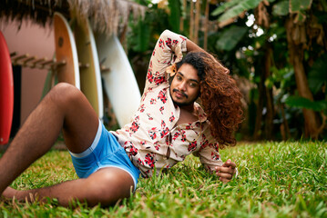 Confident gay man with rich curly hair reclines in tropical garden, vibrant style. Blue shorts,...