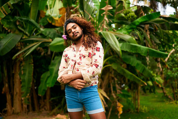 South Asian gay man with a flower in hair stands confidently in a rich garden. Wearing shorts and a...
