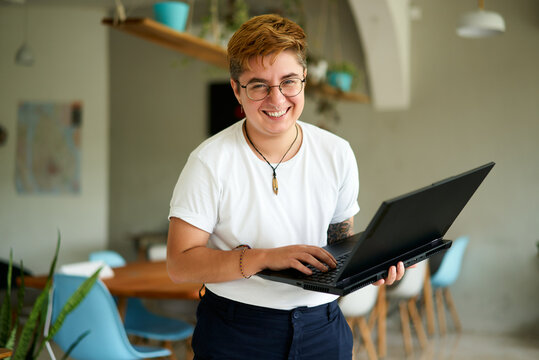 Smiling transgender professional holds laptop, works happily in modern office space. Inclusive workplace, young trans person enjoys career, experiences job fulfillment with tech in eco-friendly area. - Powered by Adobe
