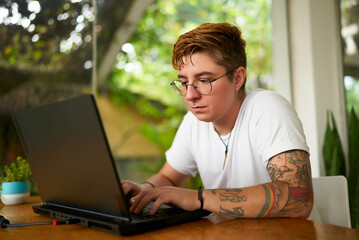 Transgender freelancer works on laptop in tropical coworking space. Focused individual with tattoos tech for remote job. Open, inclusive work plants. Creativity, productivity LGBTQ-friendly setting.