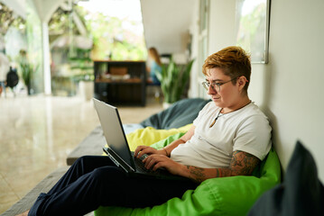 Transgender professional works on laptop, seated on green bean bag, in casual office space. Relaxed environment, light-filled room, modern work life balance, inclusion in workplace highlighted.
