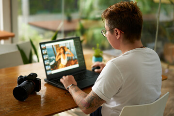 Professional photo editor works on laptop in bright coworking space. Tattooed, stylish person edits...