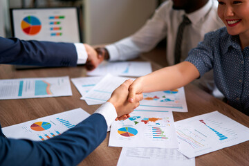 Multiracial business team seals agreement with a handshake over a desk scattered with vibrant charts, graphs, symbolizing growth, partnership, strategy, collaboration in a modern office setting.