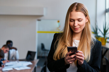 Professional blonde woman in smart casual attire uses smartphone in modern office. Focused...