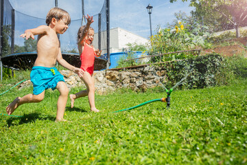 Two kids joyfully sprint through backyard sprinkler on sunny day - 784039101