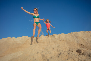 Woman and young girl leaps from sandy dune against clear sky - 784052776