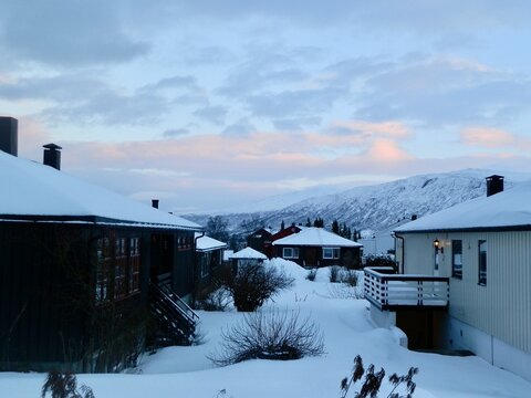 Snow-covered rooftops of a Tromso residential area at sunrise with the Tromsdalstinden mountain in the distance.