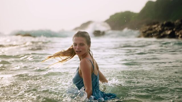 Elegant woman in blue dress enjoys ocean bath during sunset. Smiling lady relaxes in warm sea, water splashing around on romantic scenic beach. Dress flowing, serene experience in nature. Slow motion.