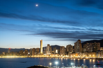 City skyline at twilight with illuminated buildings and a crescent moon above, reflecting on the...