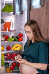 Pregnant woman near the refrigerator chooses what to eat between vegetables and dessert