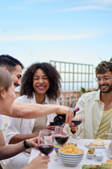Vertical. Young smiling man serving red wine to guests at food table in celebration with happy friends on rooftop. Group of cheerful multiracial friends gathered for lunch party on outdoor terrace