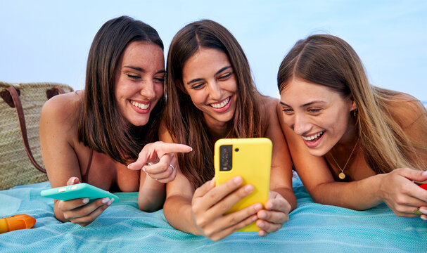 Three cheerful caucasian female friends having fun outdoors lying on a beach towel looking at one cellphone smiling together while enjoying the summer day. People addicted to technology on vacation.