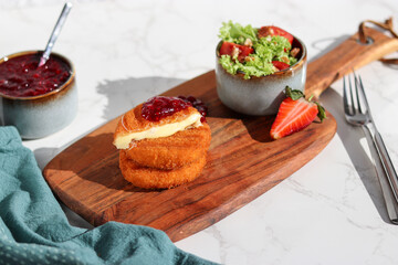 A portion of fresh made fried camembert cheese with cranberry sauce on top and a side of fresh salad on wooden plate