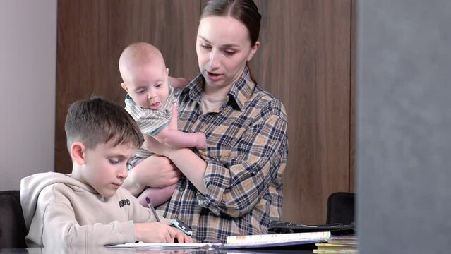 Mother with a baby helps her eldest son do his homework. Education, family lifestyle, homeschooling concept.