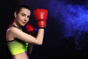 Portrait of beautiful woman wearing boxing gloves in color lights and smoke on black background. Space for text
