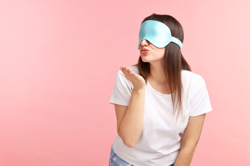 Woman in pyjama and sleep mask blowing kiss on pink background, space for text