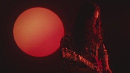 Rock musician with long curly hair playing electric guitar during performance in dark studio with red light projection