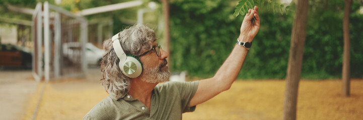 Panorama, Friendly middle-aged man with gray hair and beard wearing casual clothes listening to music on headphones. Mature gentleman in eyeglasses enjoys music outdoors