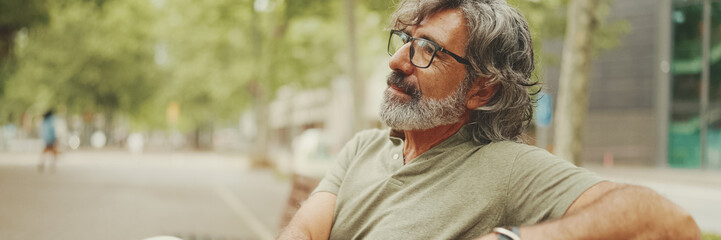 Positive middle-aged man with gray hair and beard wearing casual clothes sits on bench, Panorama. Mature gentleman in eyeglasses is resting on bench