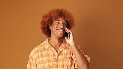 Smiling stylish curly guy speaks emotionally on mobile phone, isolated on brown background in the...
