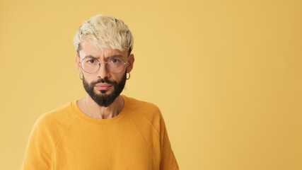 Guy with glasses, dressed in yellow T-shirt, listens carefully and strongly disagrees, isolated on yellow background in studio