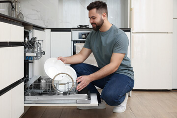 Smiling man loading dishwasher with plates in kitchen