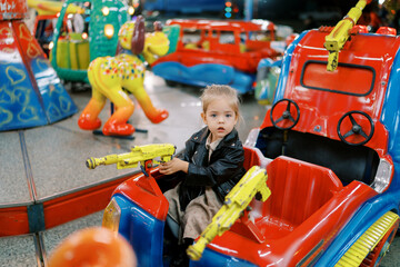 Little girl sits in a toy car on a carousel, holding the steering wheel-gun