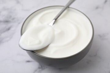 Delicious natural yogurt in bowl and spoon on white marble table, closeup