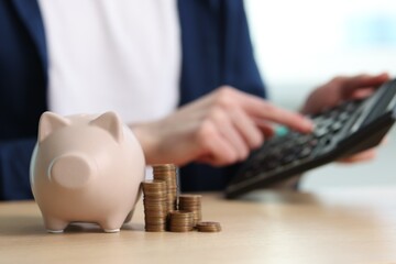 Financial savings. Man using calculator at wooden table, focus on piggy bank and stacked coins - obrazy, fototapety, plakaty