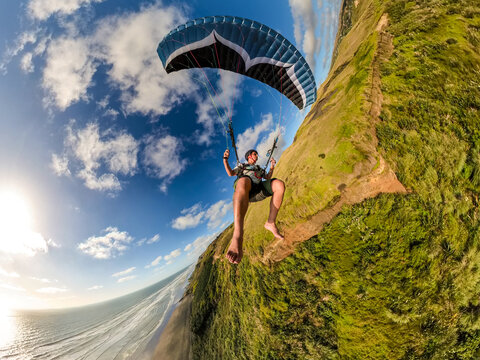 Extreme paragliding pilot soaring along a New Zealand beach at sunset, flying barefoot in warm summer light